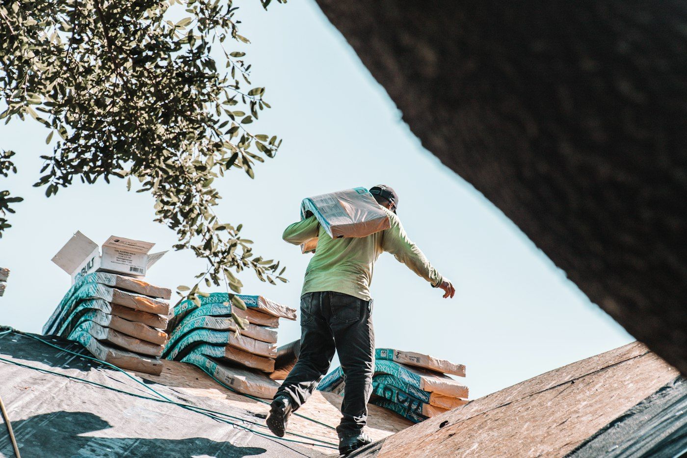 Roofer carrying a bundle of shingles across a papered roof deck, seen from behind
