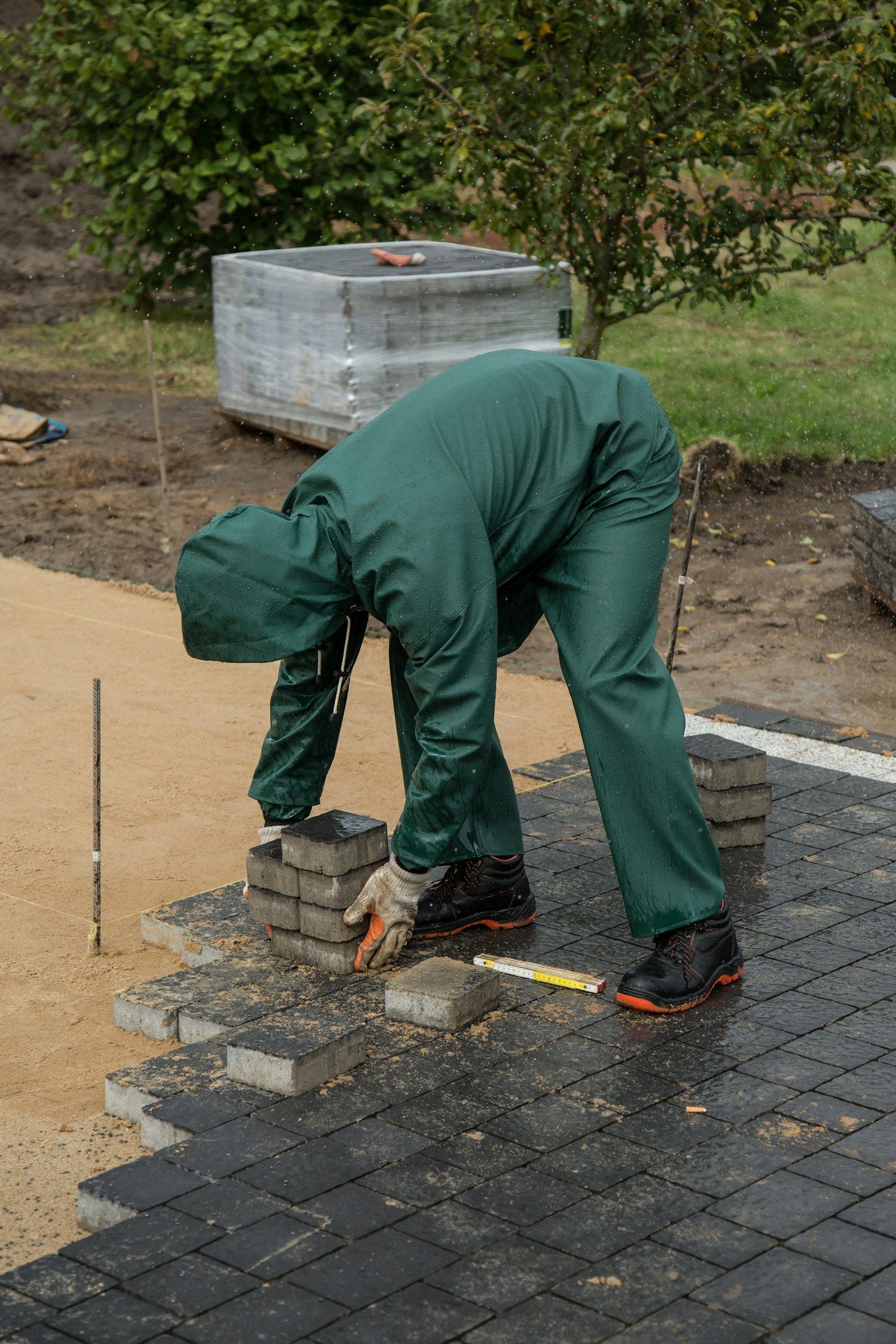 Installer in green coveralls bending to set pavers on a screeded sand bed