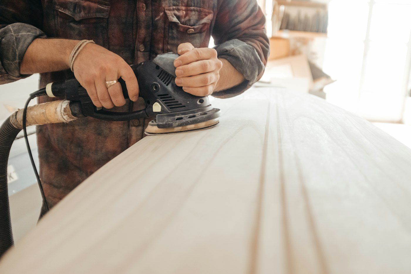 Hands guiding a random orbit sander with a dust hose across a pale board