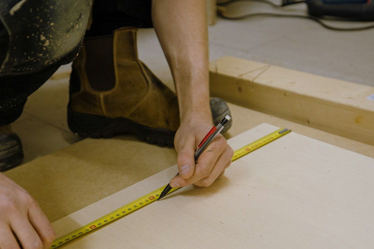 Installer kneeling on the subfloor, marking a line on a plywood panel with a tape and pencil