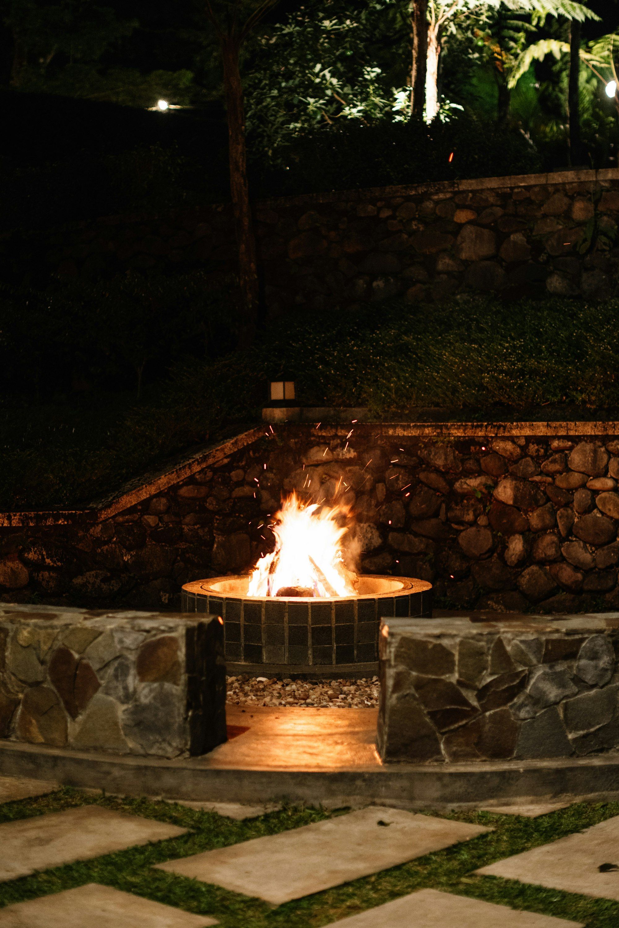 Stone fire pit lit at night with a fieldstone wall behind and square pavers set in grass