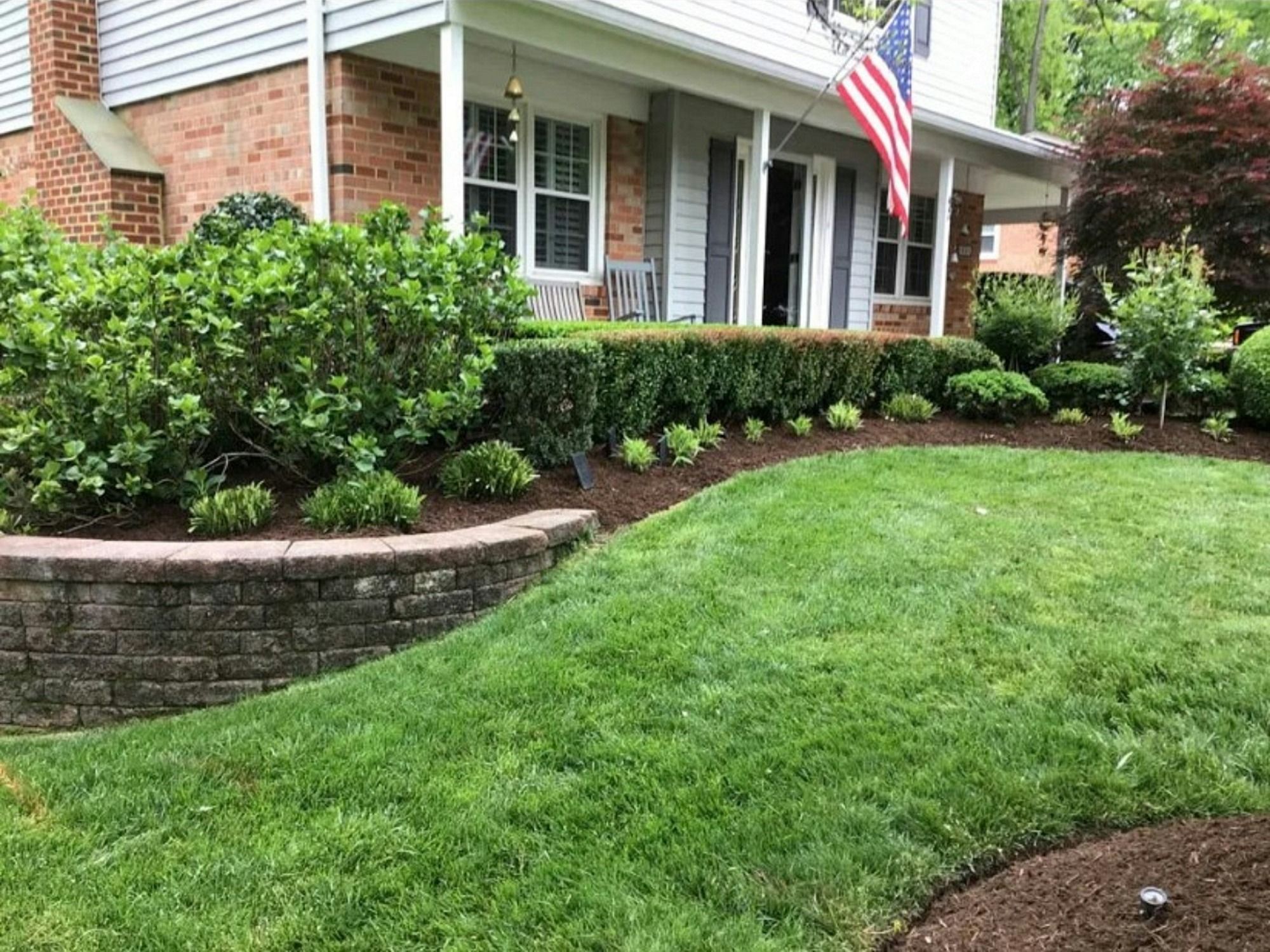 Curved block retaining wall holding a mulched shrub bed in front of a brick colonial