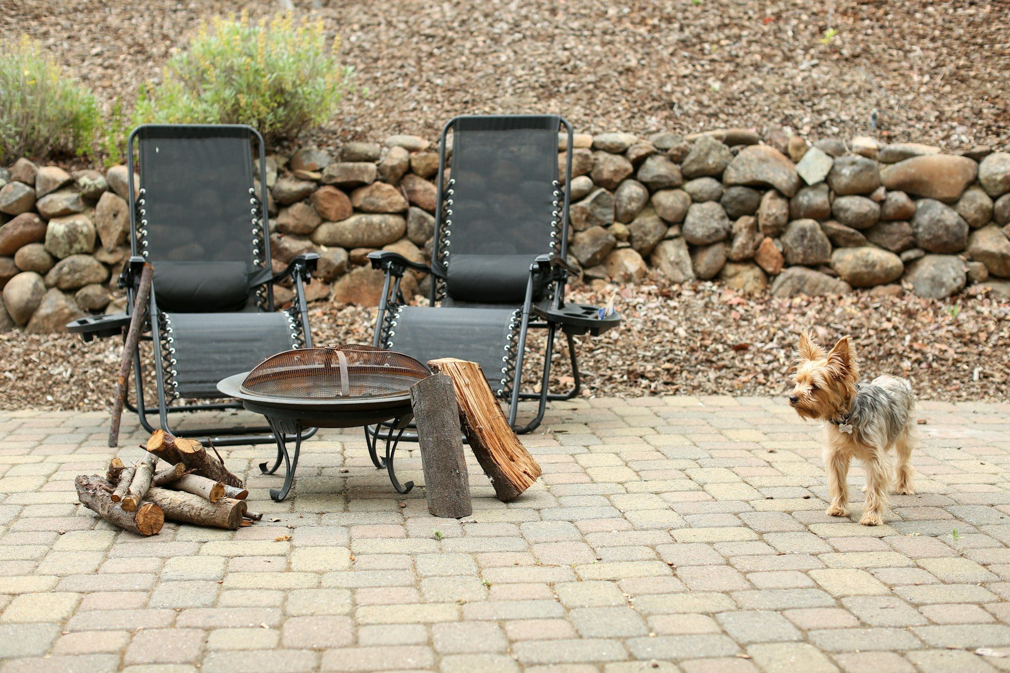 Tan paver patio with lounge chairs and a fire bowl against a round-stone wall