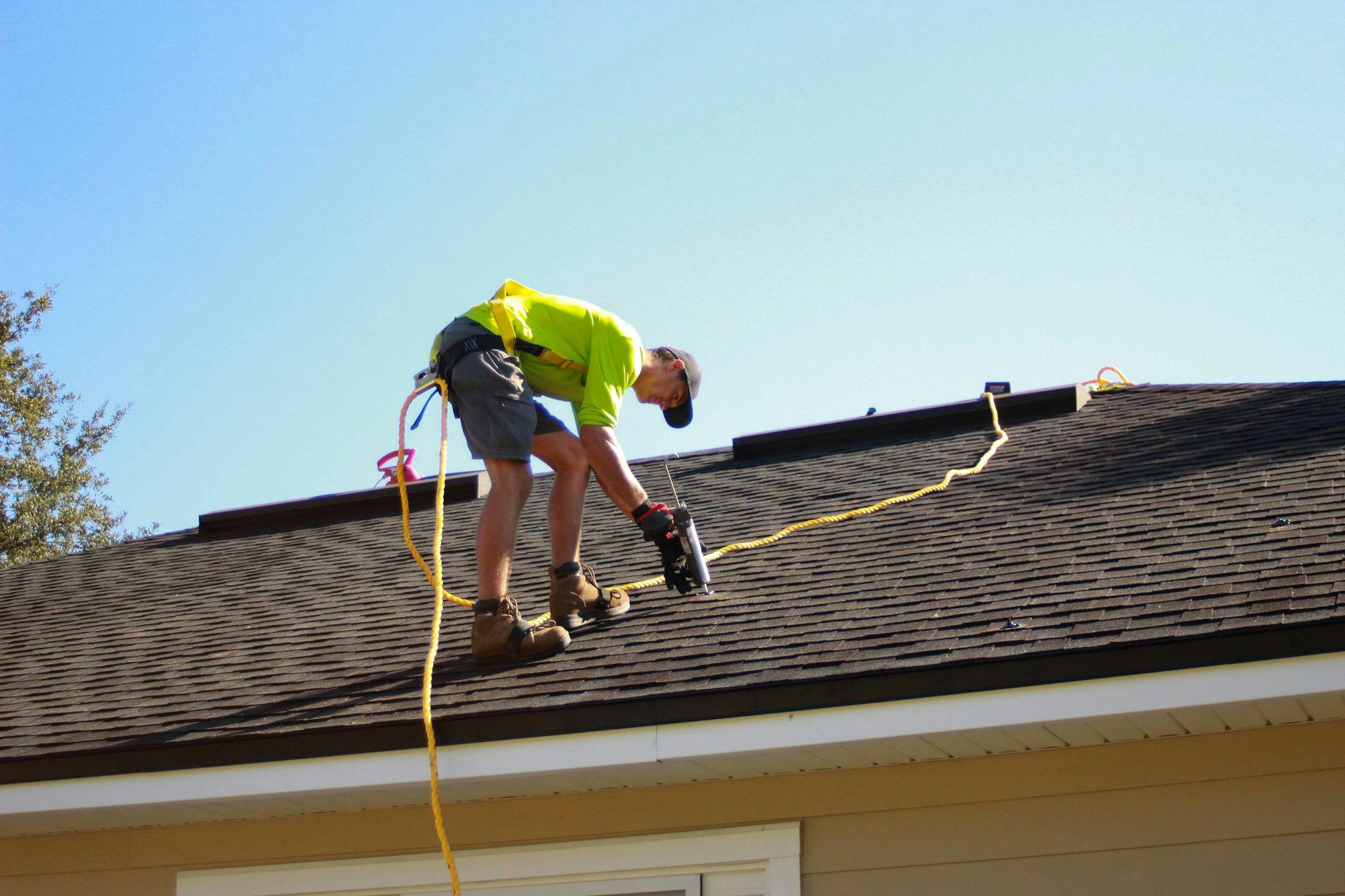 Ridgeline roofer in a hi-vis shirt and fall-protection harness sealing exposed nail heads on dark architectural shingles