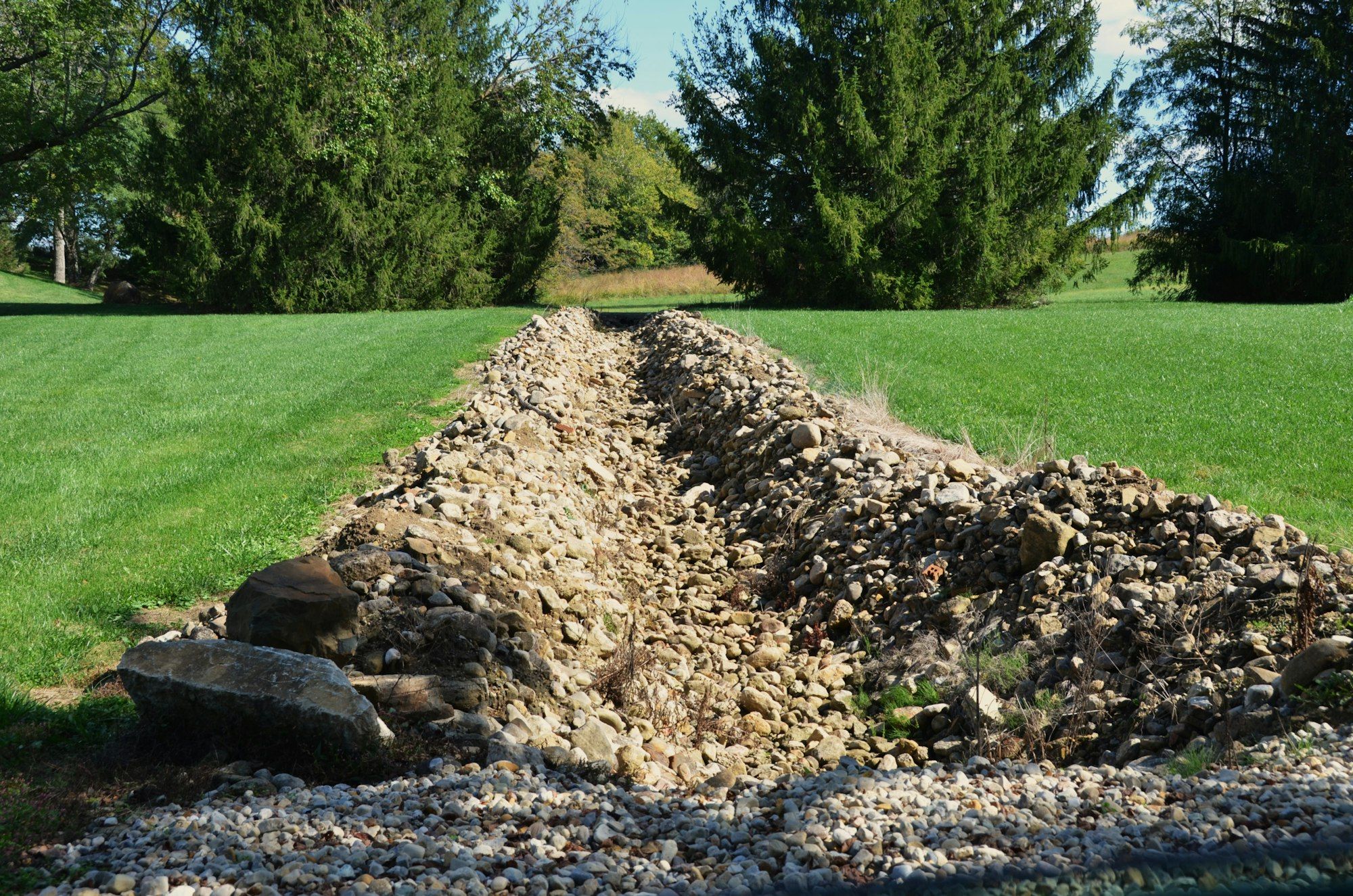 Rock-lined drainage swale running down a sloped lawn toward a row of evergreens